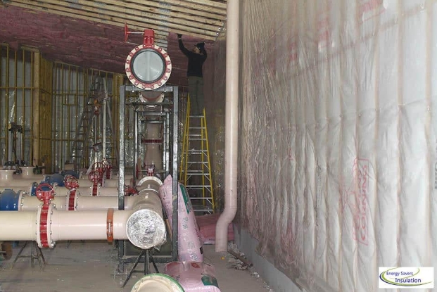 A man is standing on a ladder in a room filled with pipes.