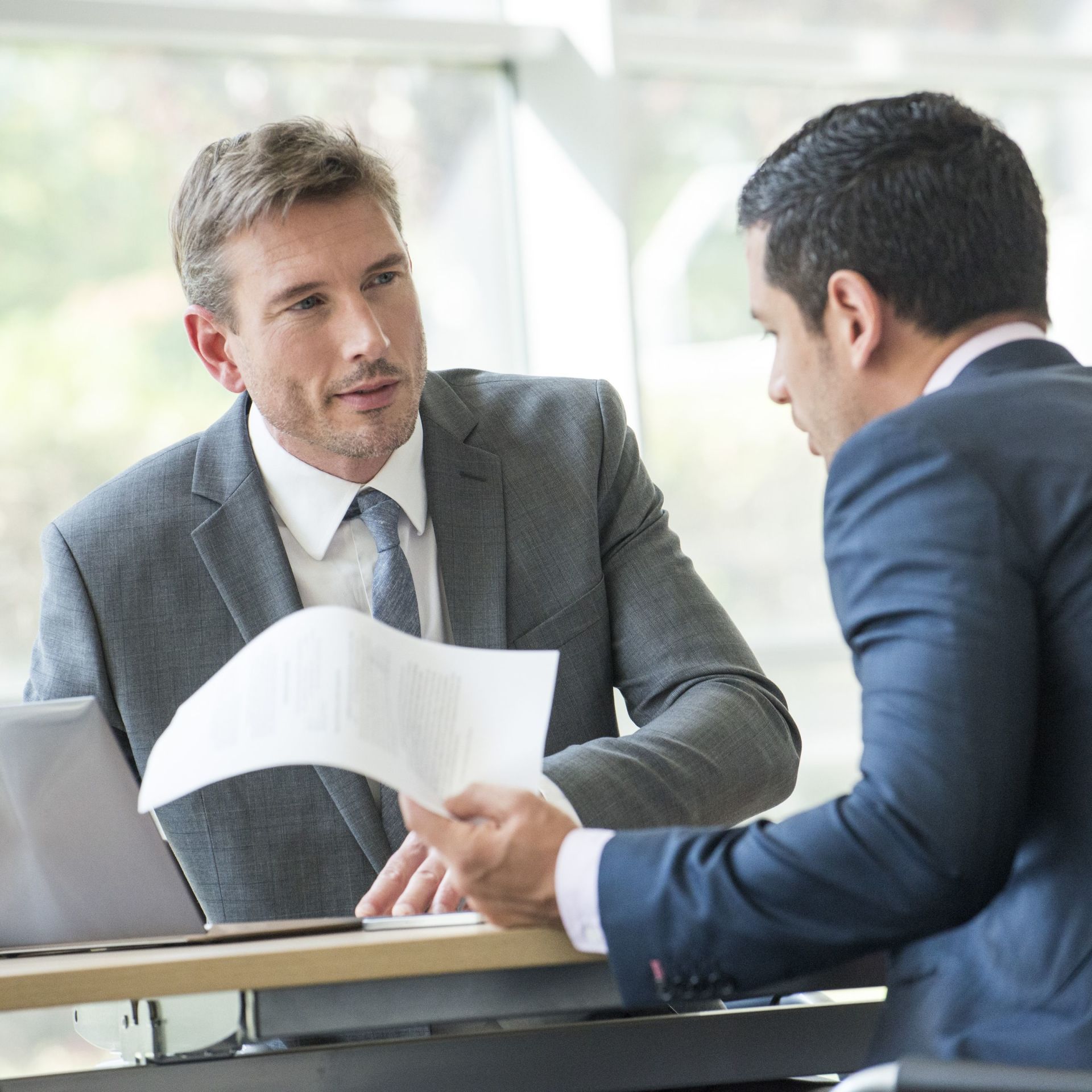 Two men in suits at a desk, reviewing documents in an office setting.