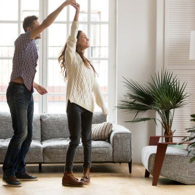 A couple dancing in a bright living room, man raises woman's hand. Gray couch, potted plant visible.