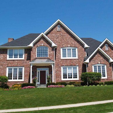 Brick two-story house with white trim, dark roof, and lush green lawn against a clear blue sky.