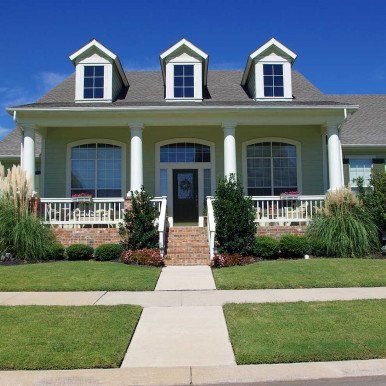 Green house with white columns, brick steps, and three dormers under a clear blue sky.