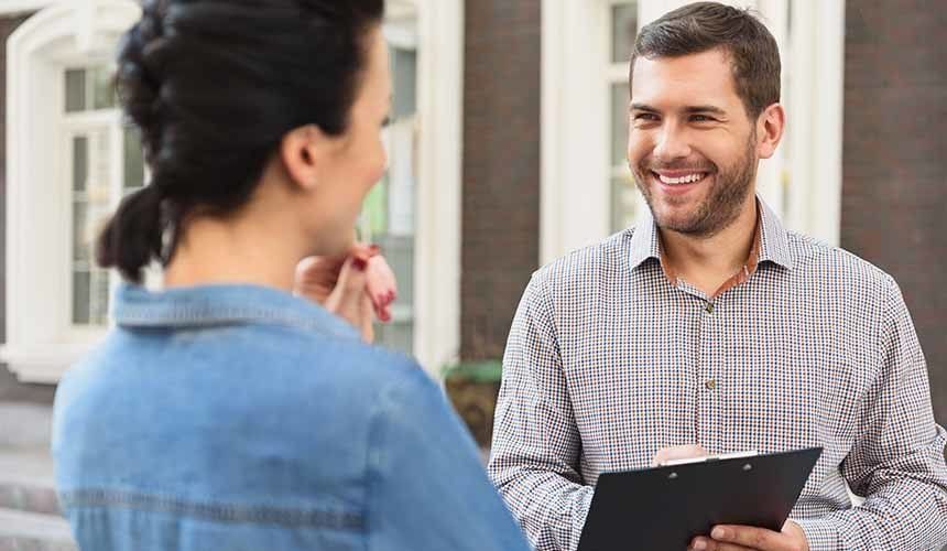 Man in plaid shirt smiles, holding clipboard, talking to a woman in front of a house.