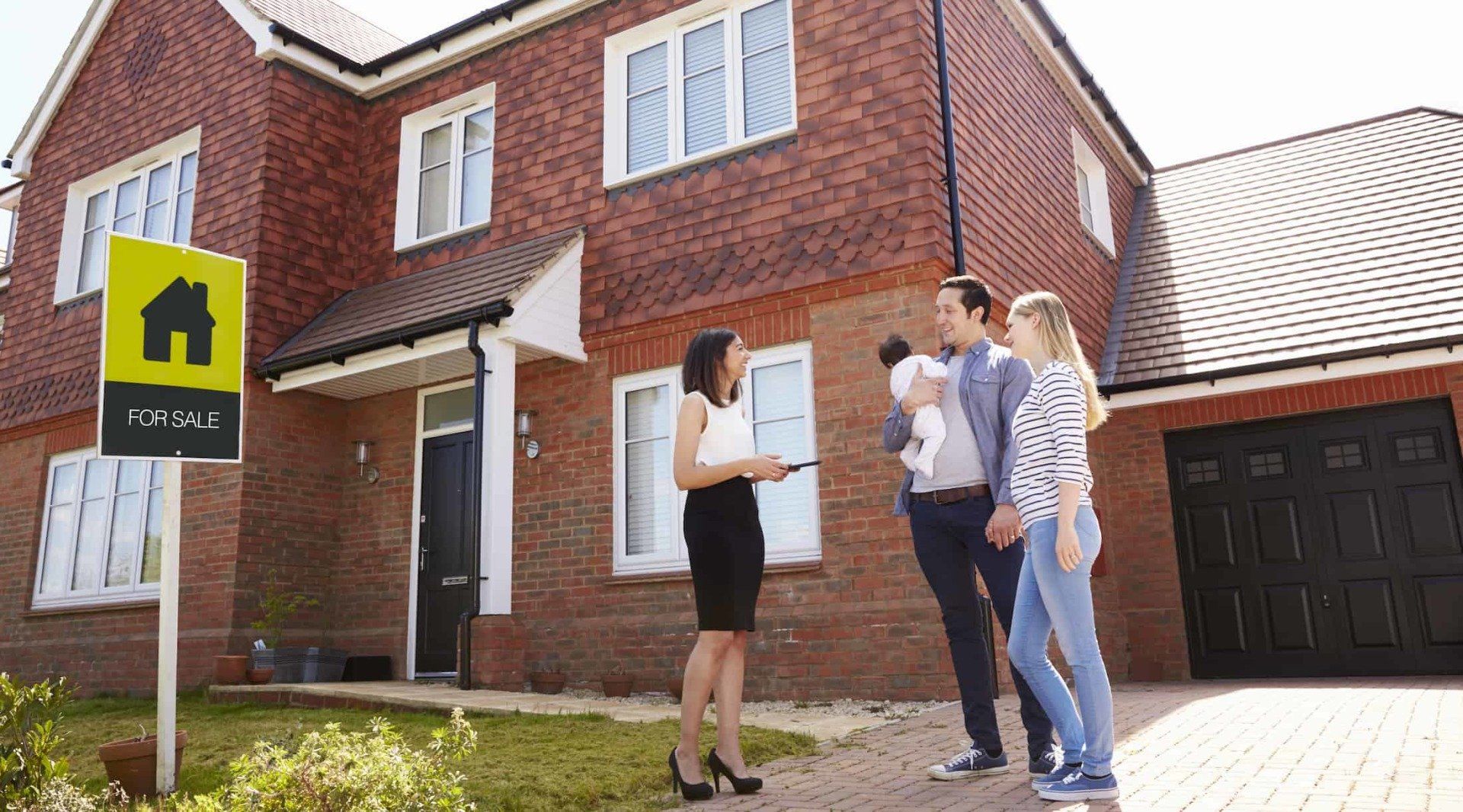 Real estate agent showing house to a couple with a baby. “For Sale” sign in front.