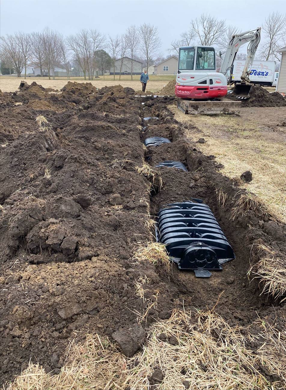 Construction site with a backhoe excavating a trench; a black plastic septic tank is in the trench.