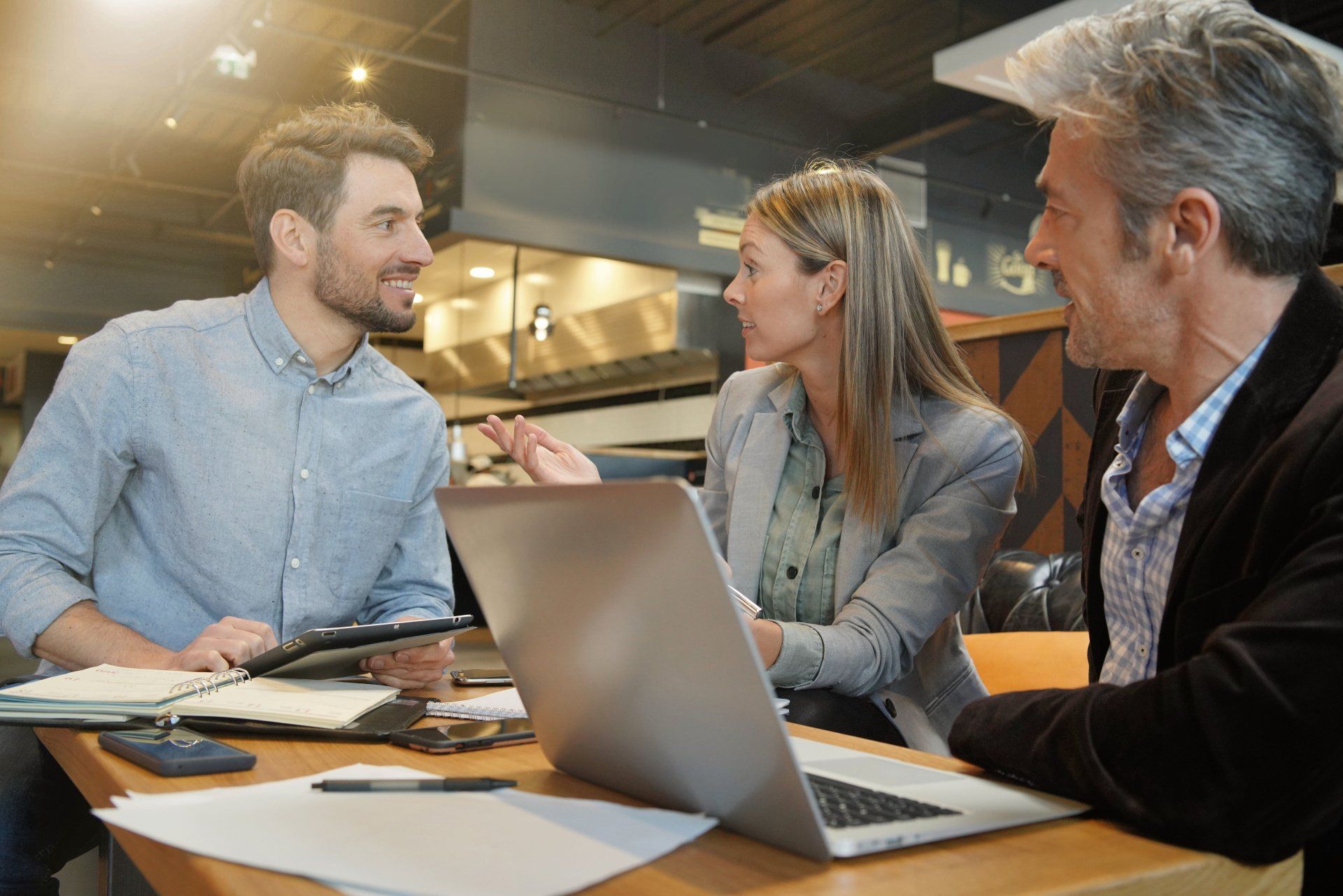 Three professionals in a modern office space discuss a project around a table with a laptop and documents.