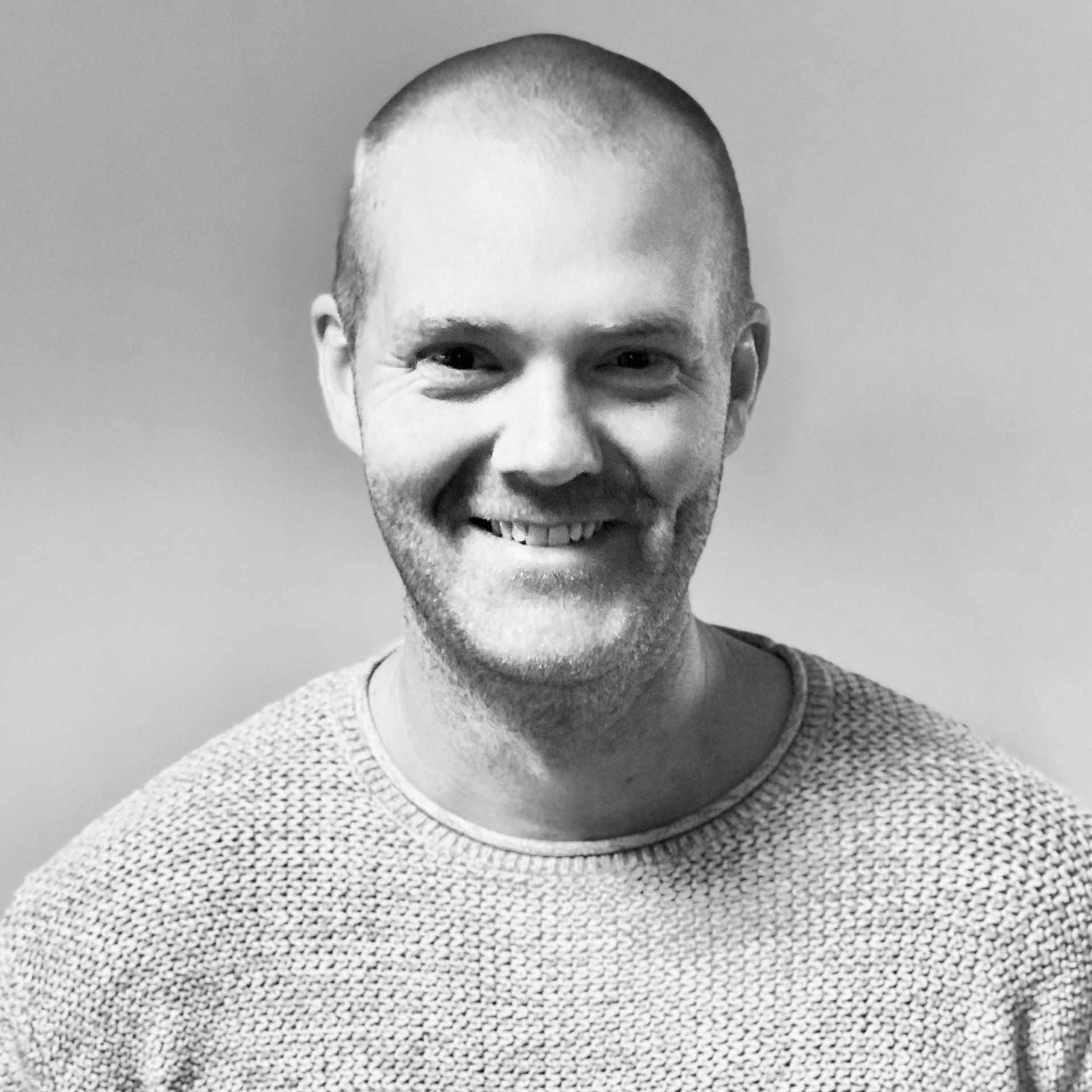 A headshot of a person with a short haircut, wearing a textured sweater, smiling warmly at the camera against a gray wall.