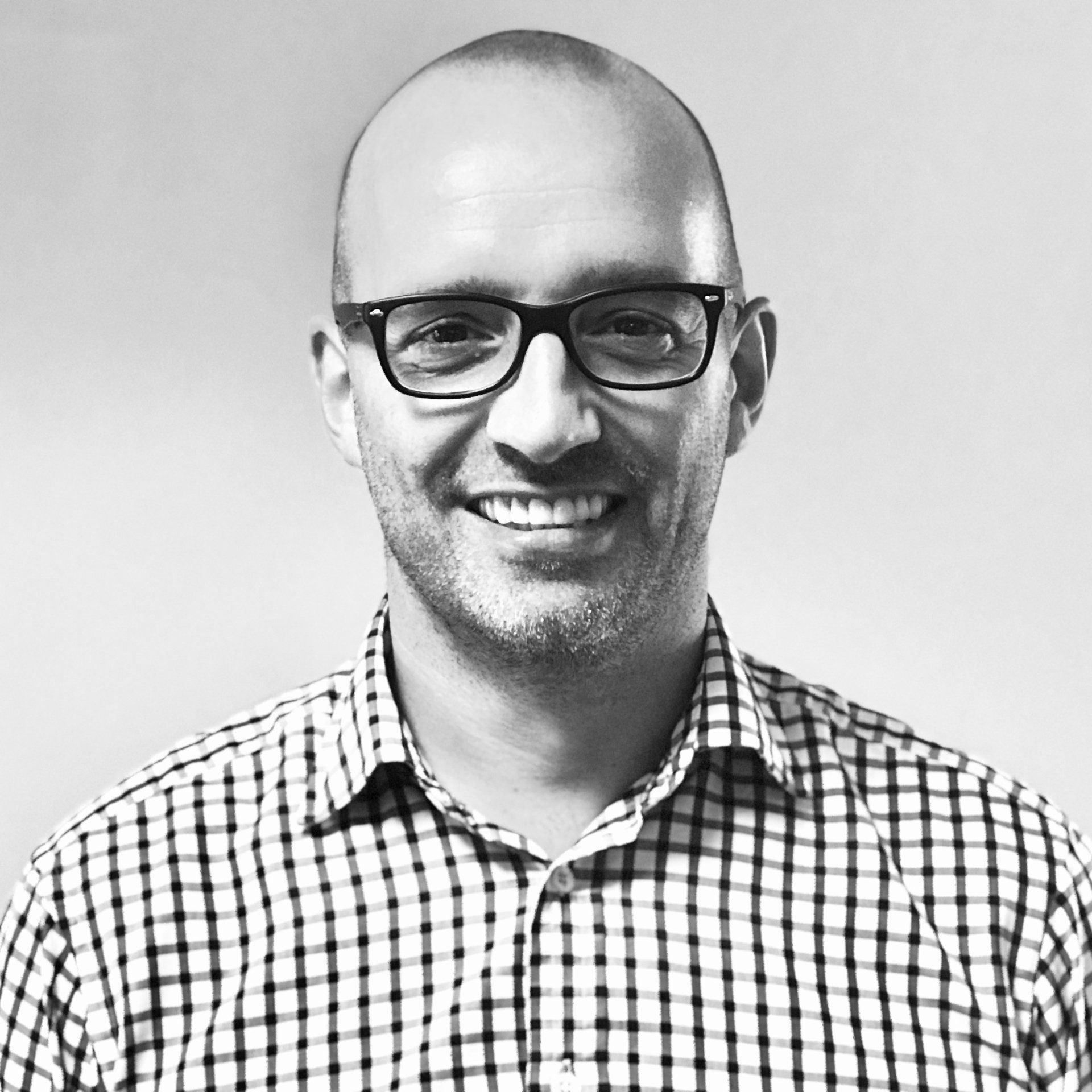 A smiling man with a shaved head and glasses, wearing a checkered button-down shirt against a plain, light background.