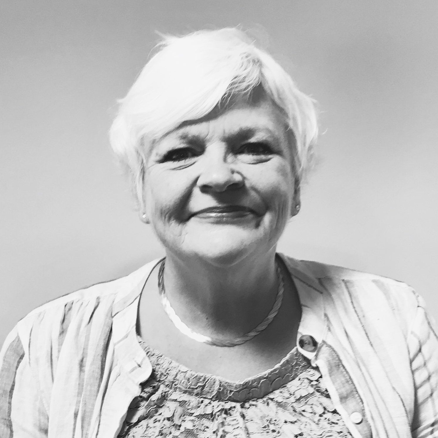 A smiling person with short, light-colored hair, wearing a layered top and a necklace against a plain background.