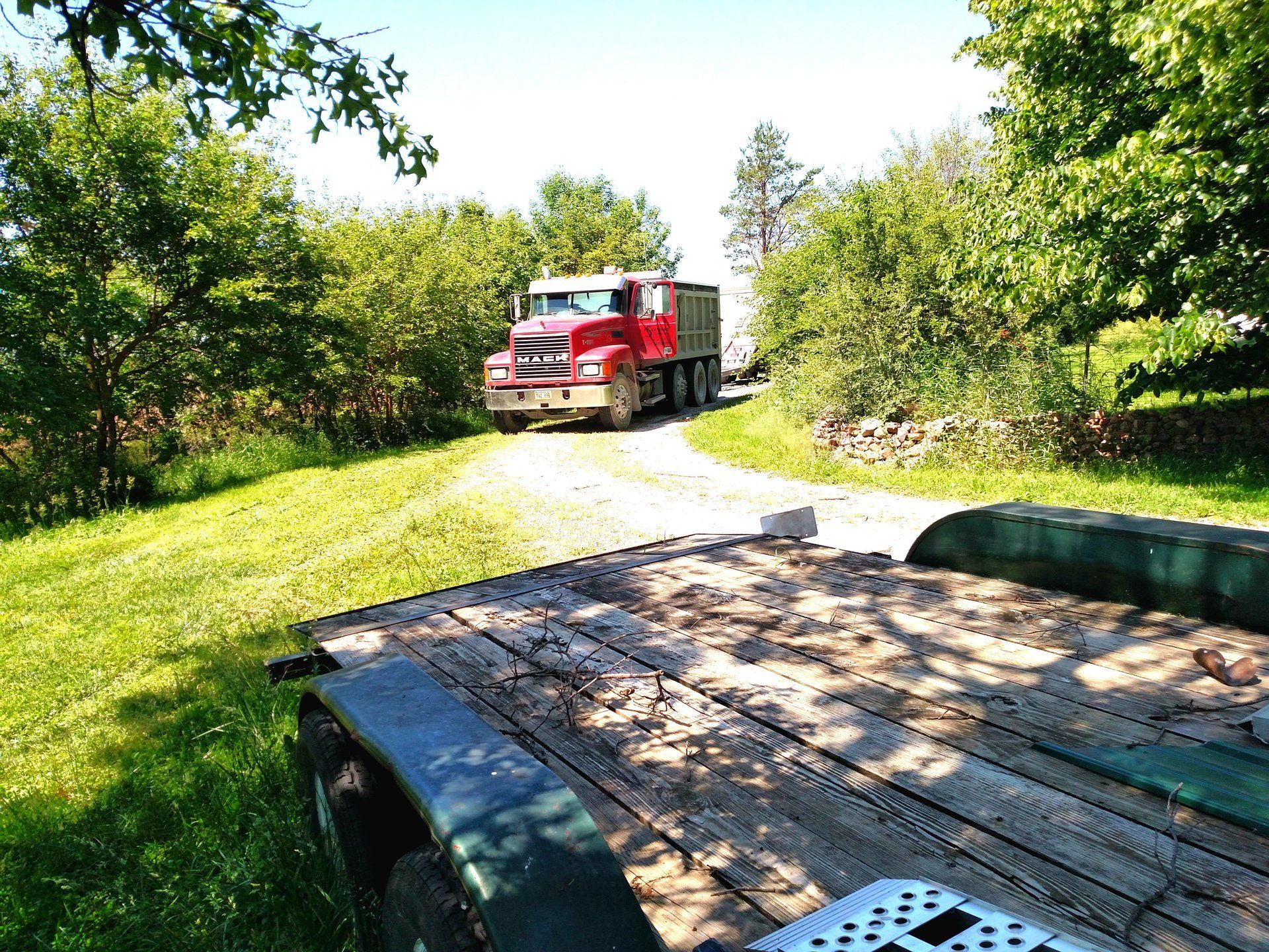 Red dump truck on gravel road, trailer in foreground. Lush green trees on either side, sunny day.