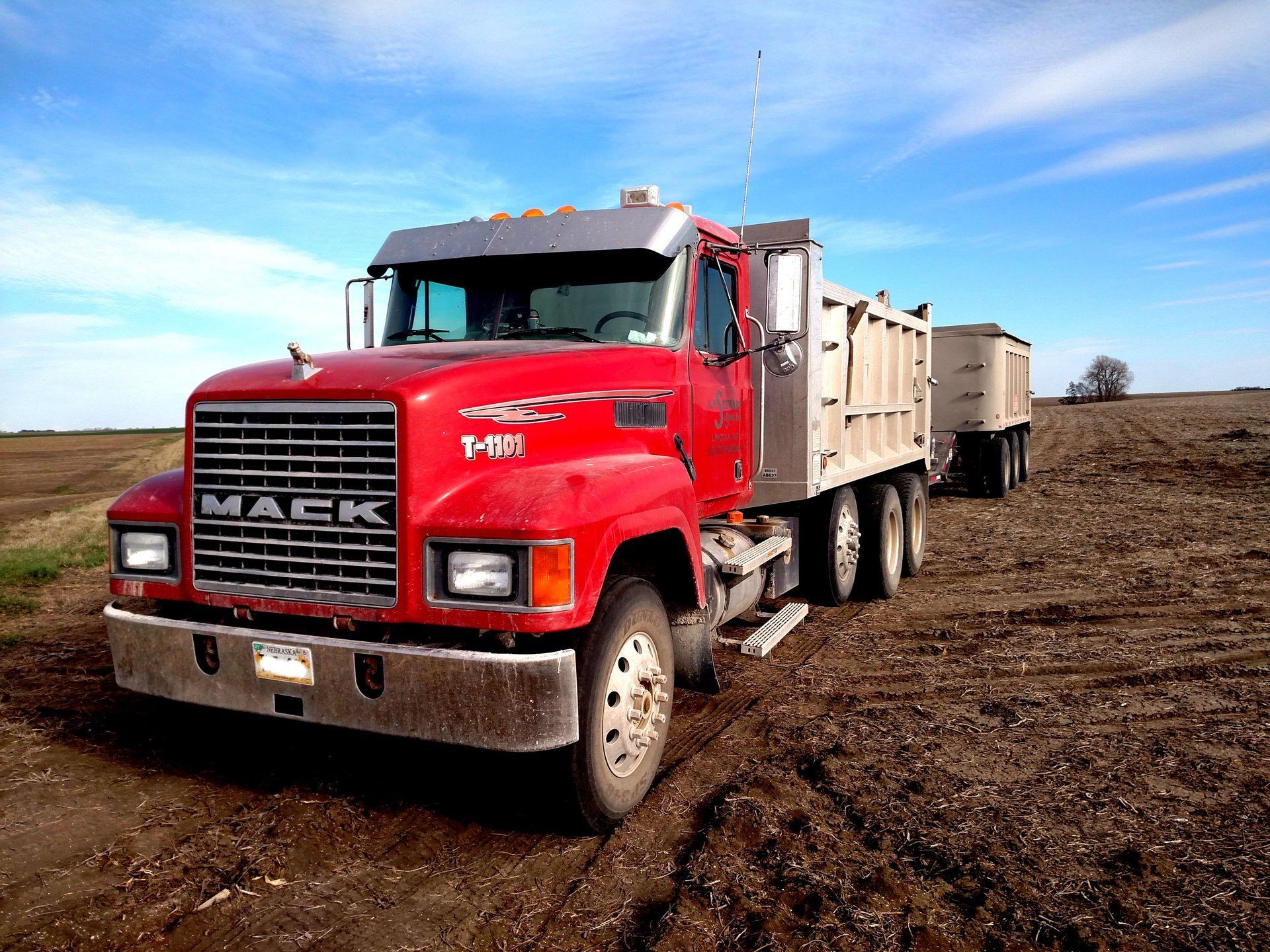 Red Mack truck hauling a trailer on a dirt field under a blue sky.
