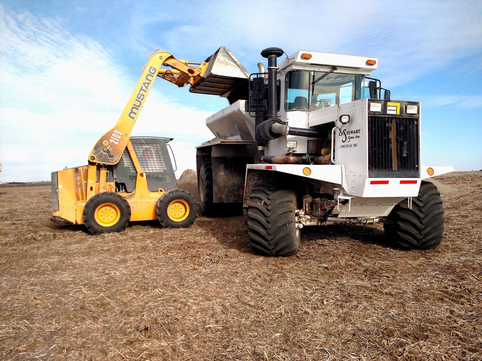 Yellow skid steer loading debris into a large white and black grinder on a brown field.