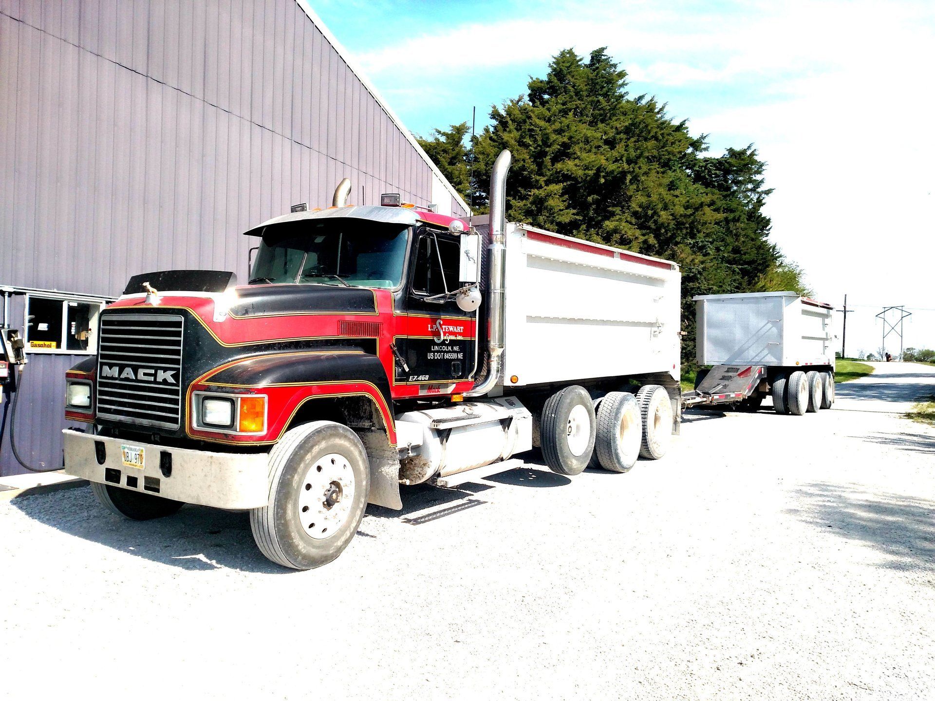 Red and black Mack truck with a dump trailer parked outside a building on a sunny day.