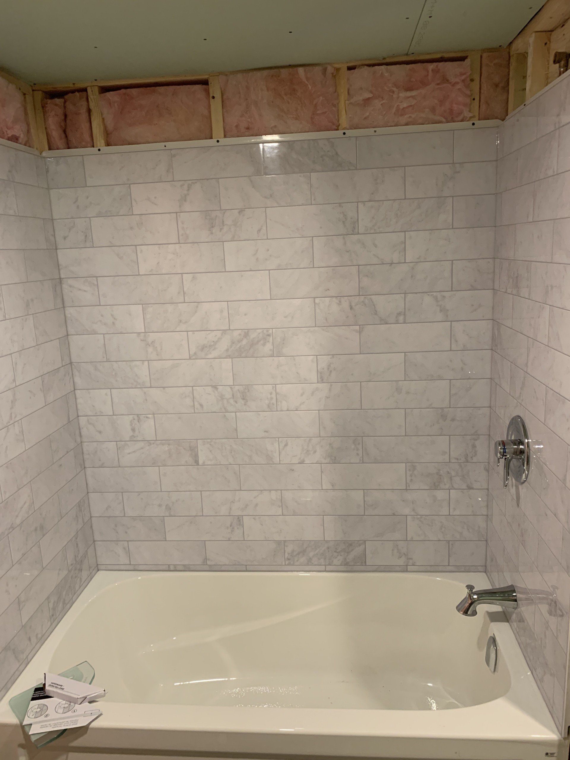 White tiled shower surround above a bathtub, with exposed framing and pink insulation visible.