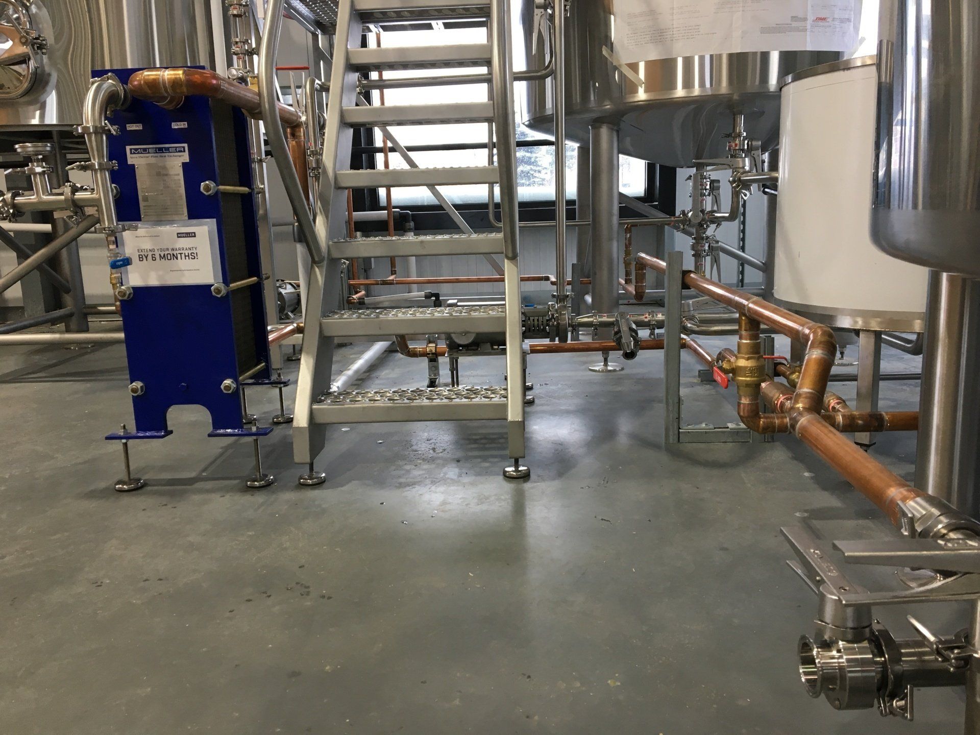 Brewery interior with stainless steel tanks, copper piping, a blue heat exchanger, and metal stairs.