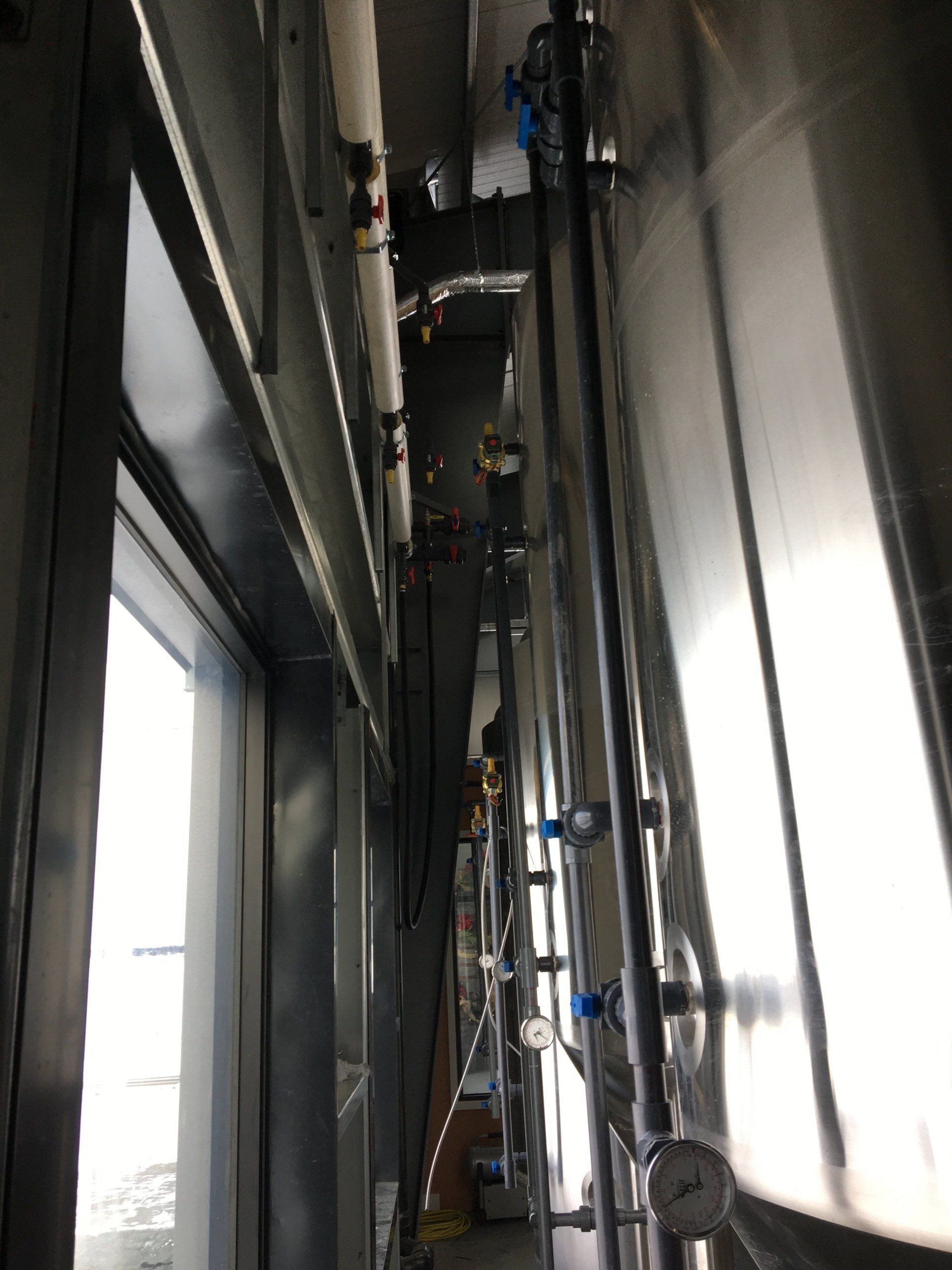 Pipes and gauges run alongside a stainless steel tank, framed by a window and metal structure.