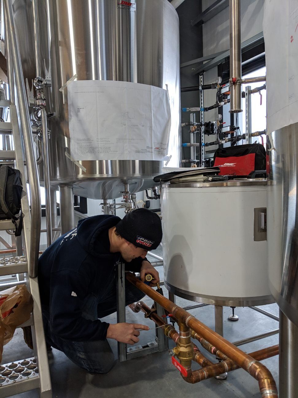 Man inspecting copper pipes in a brewery, pointing; stainless steel tanks in background.