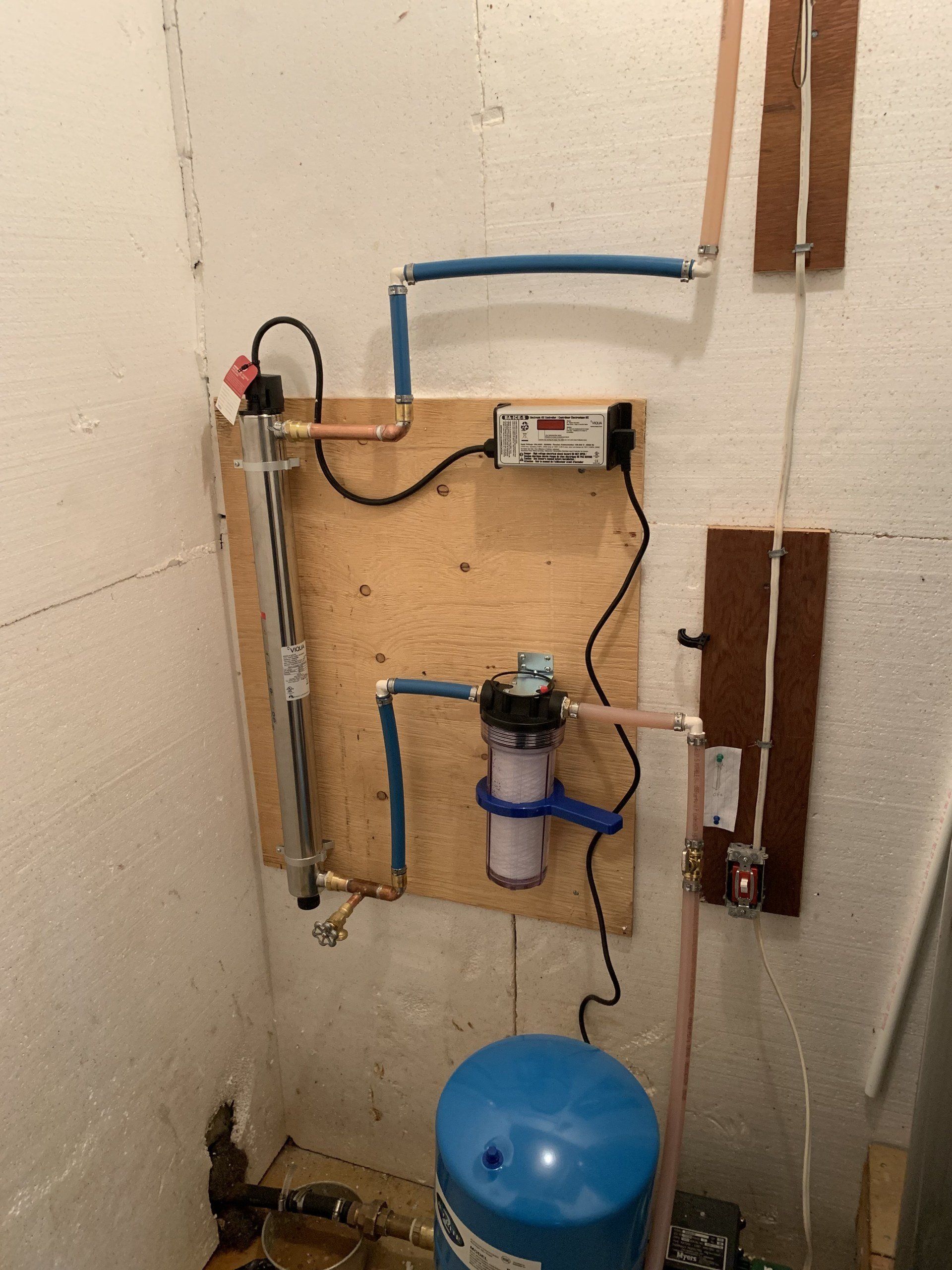 Water filtration system mounted on plywood in a white-walled room, copper pipes, blue tank, and filter visible.