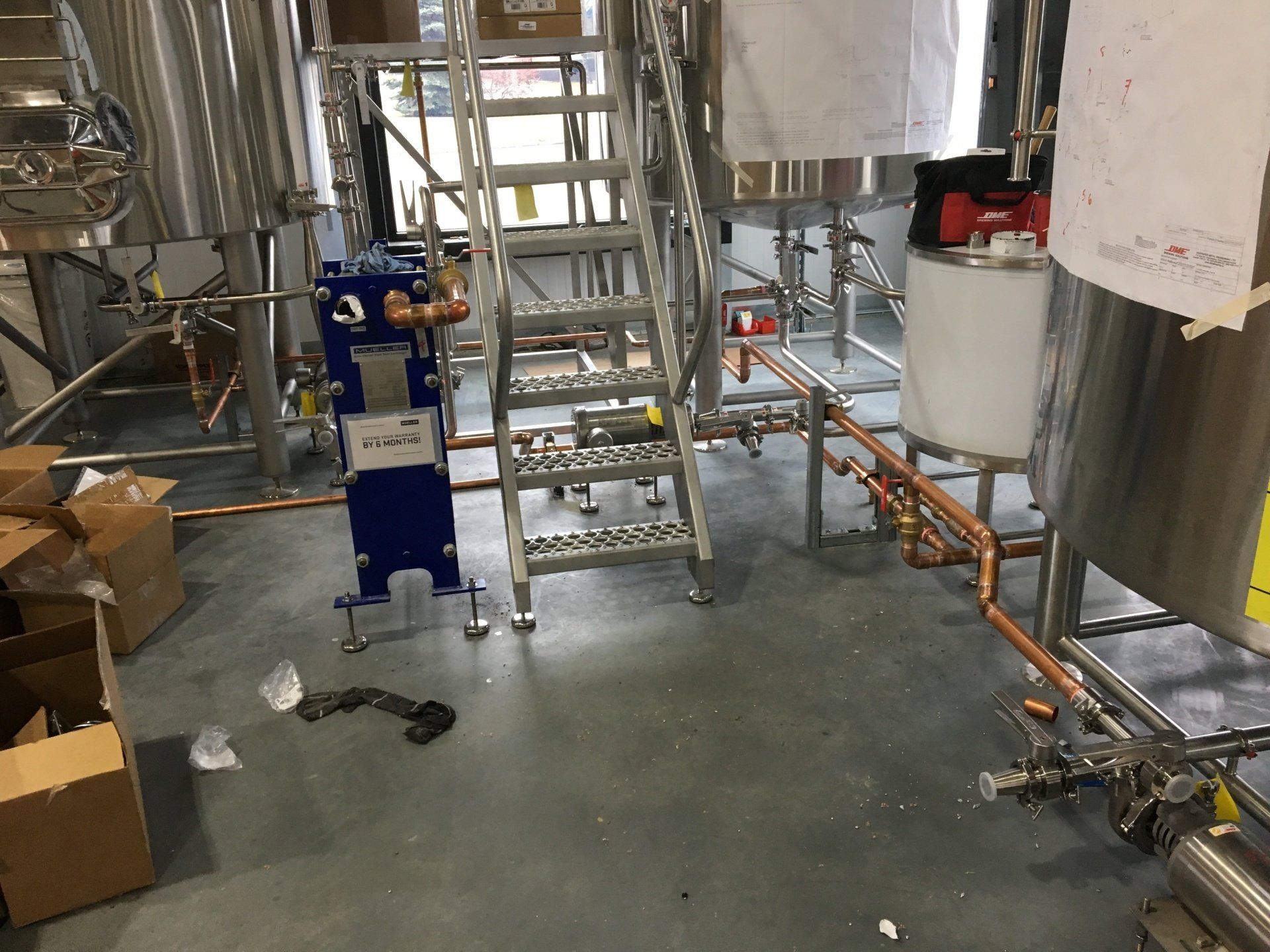 Brewery equipment with stainless steel tanks, copper pipes, and a ladder on a concrete floor.