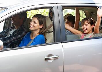 Family riding in a silver car, with a smiling passenger and a child waving in the back seat