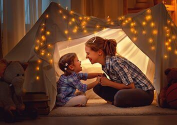 Woman and child sitting in a cozy blanket fort lit by warm string lights, facing each other and smiling.