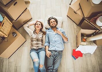 Two people lying on the floor among moving boxes, smiling and holding hands