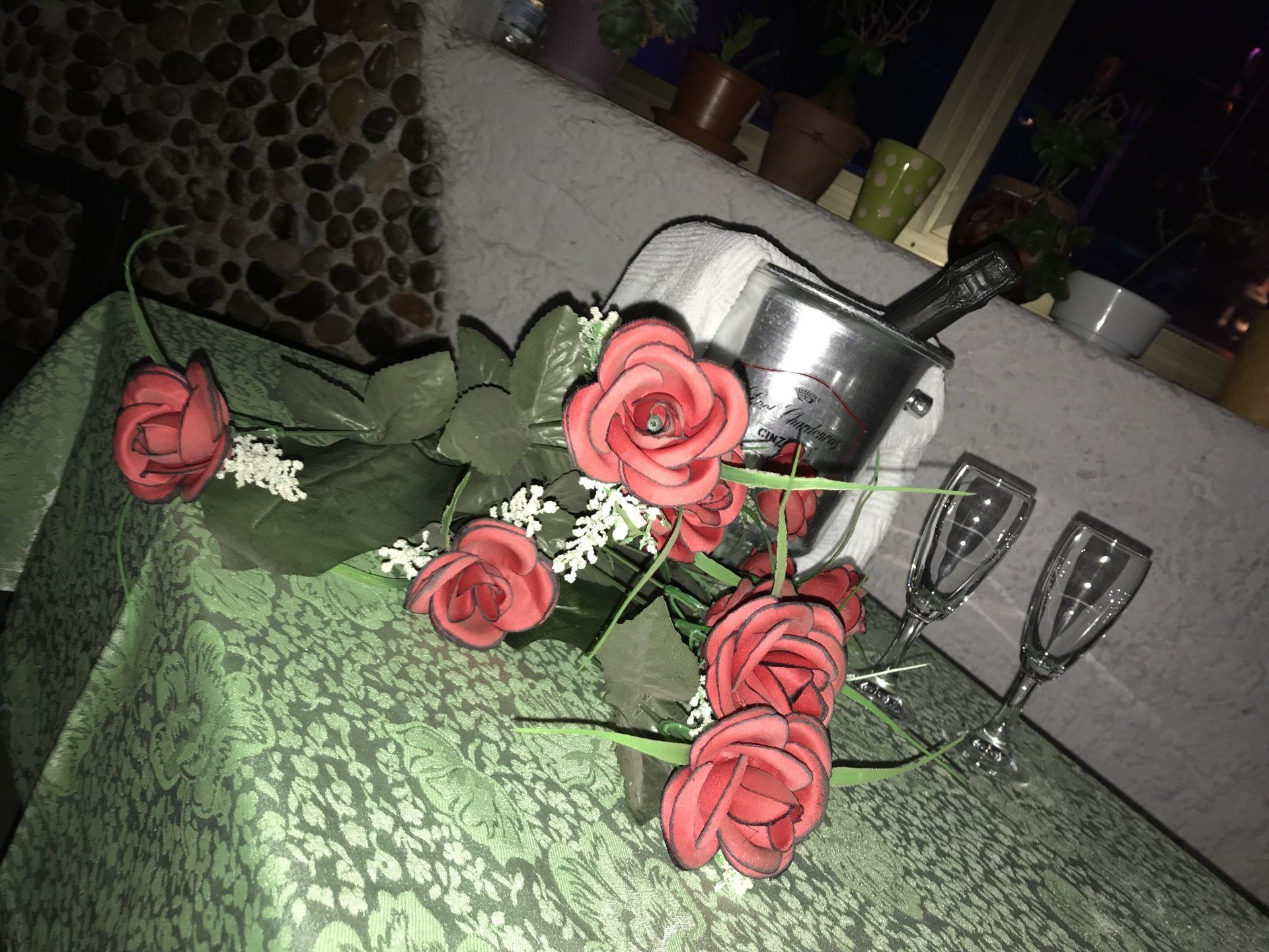 Rose artificiali rosse poggiano su una tovaglia verde a motivi geometrici, accanto a un secchiello per il ghiaccio in metallo con una bottiglia e due bicchieri alti.
