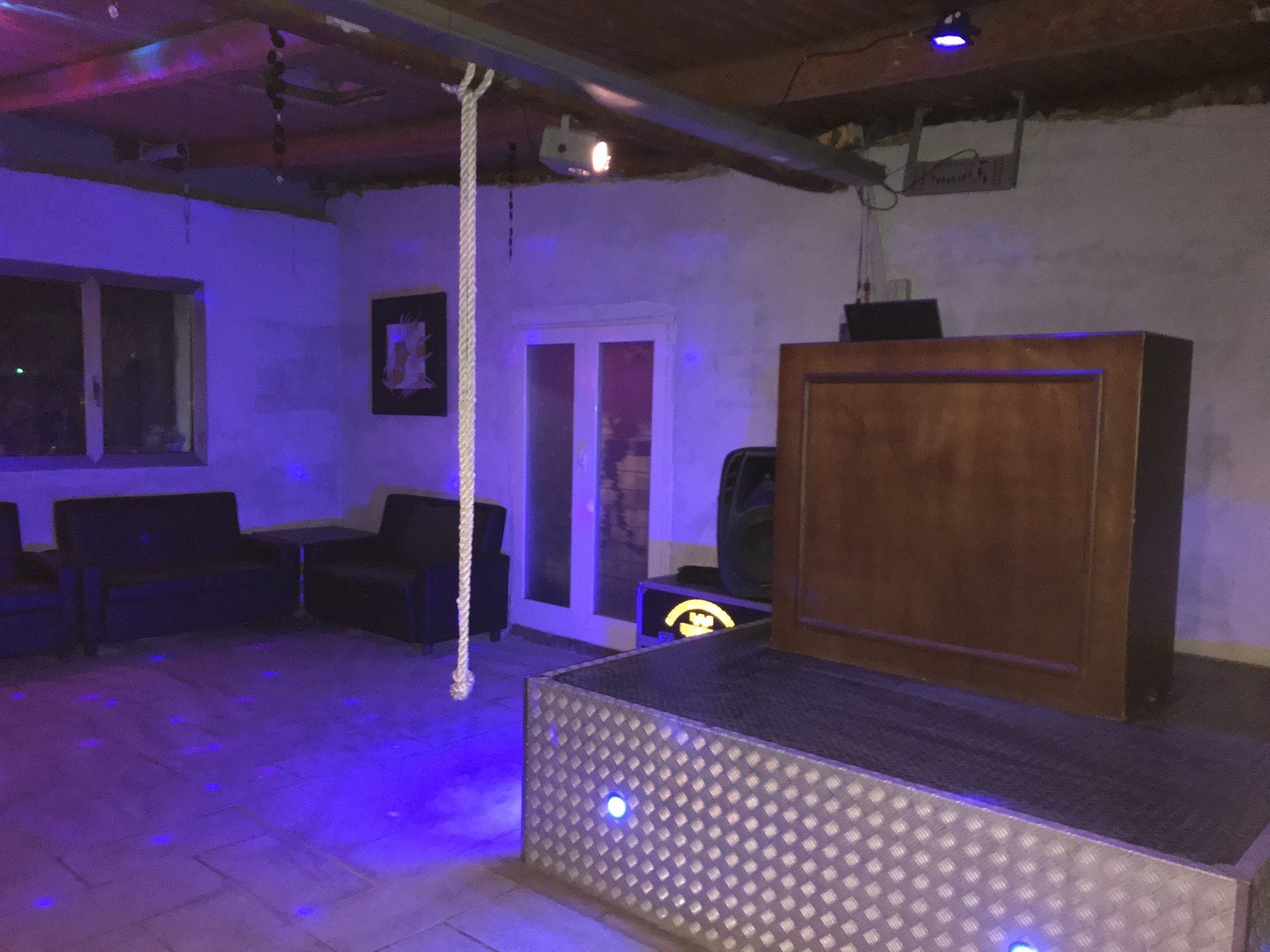 Una stanza scarsamente illuminata da luci viola, con una postazione da DJ su un palco metallico, sedie nere e una corda bianca sospesa.