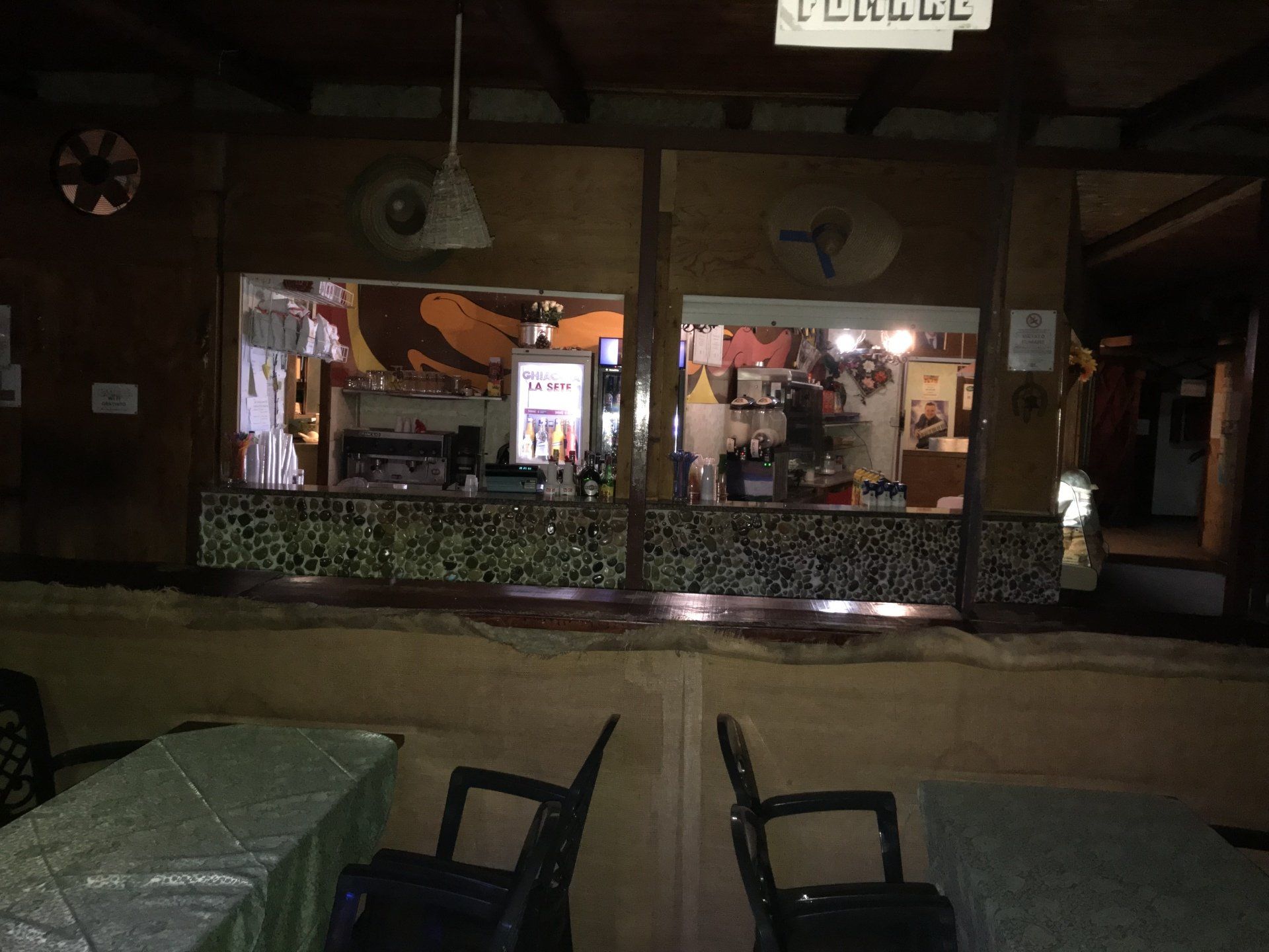 Uno spazio interno scarsamente illuminato con due tavoli e sedie disposti di fronte a un bancone di servizio con un frigorifero espositore illuminato.