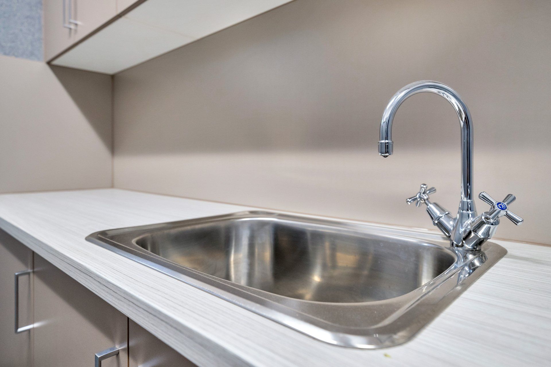 Stainless steel sink with chrome faucet on a white countertop in a neutral-toned room.