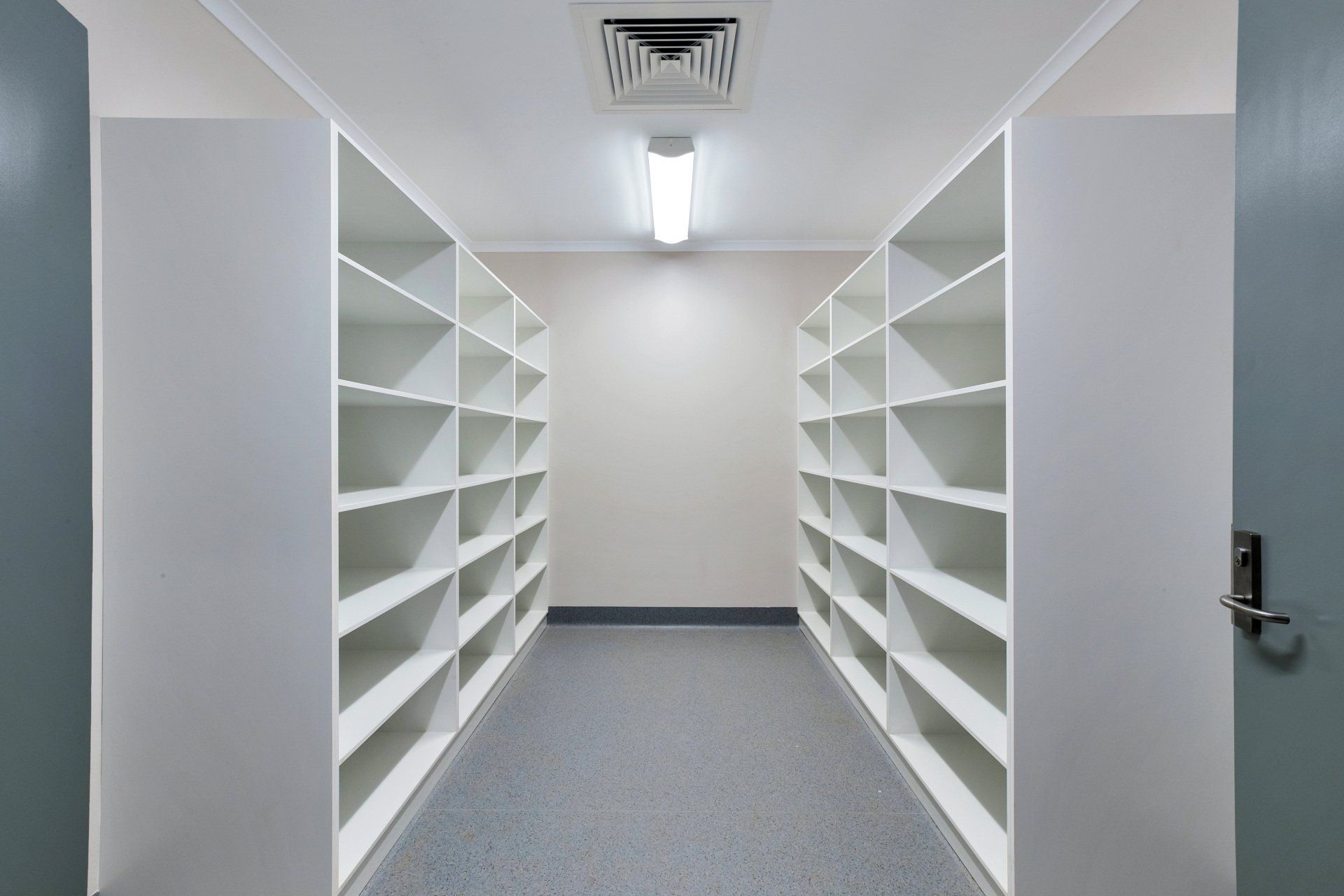 Empty storage room with white shelves on each side, blue flooring, and a bright overhead light.