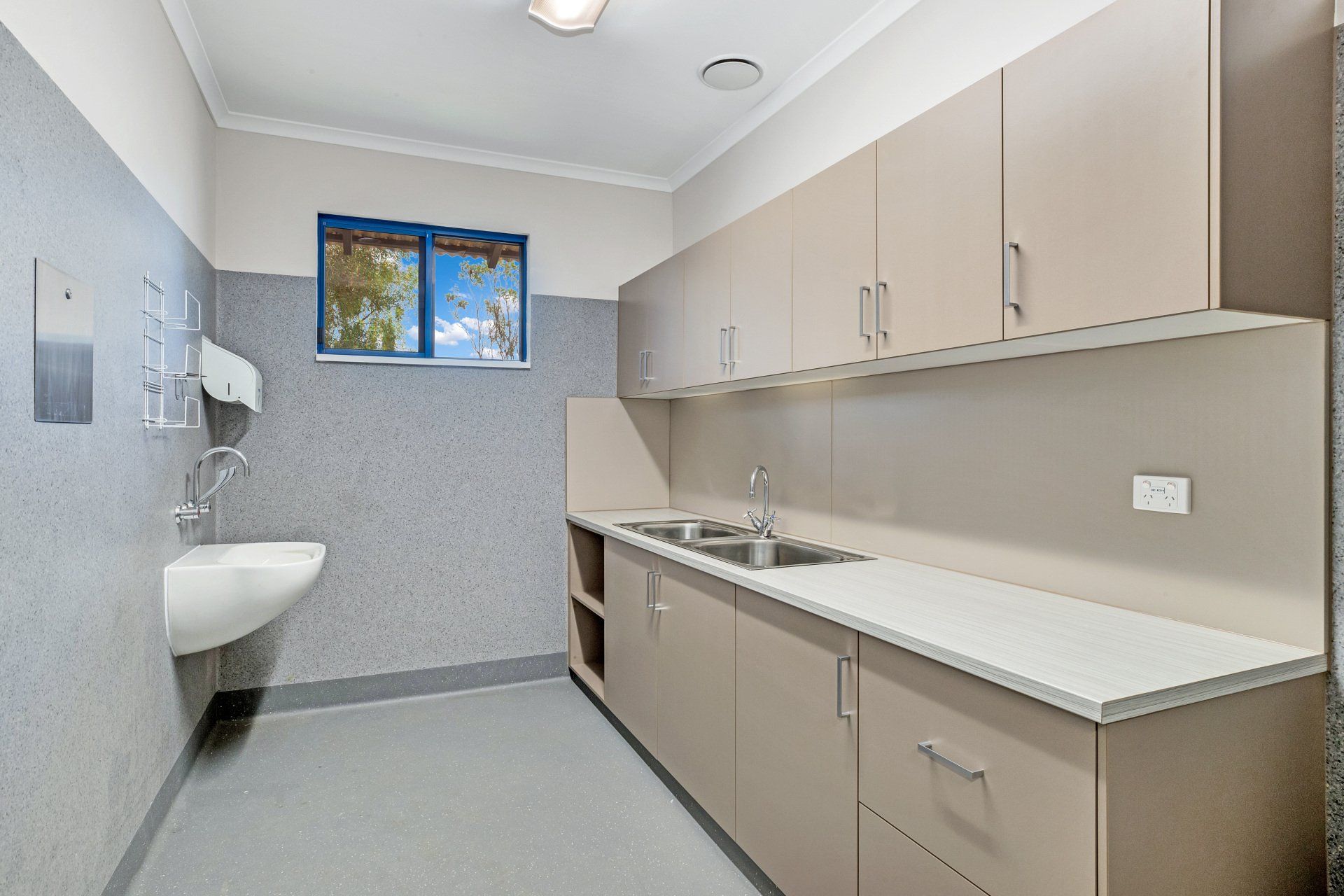 Clean, light-colored medical washroom. Gray walls, tan cabinets, white sink, blue-sky window.