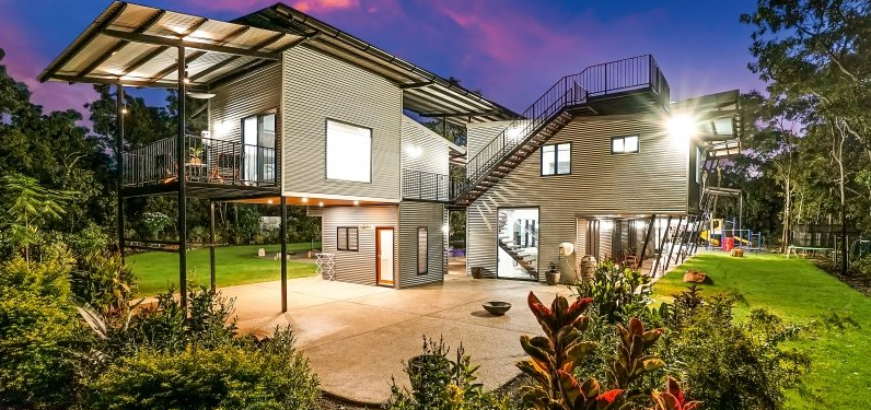 Modern home built with stacked shipping containers, surrounded by lush greenery at dusk.
