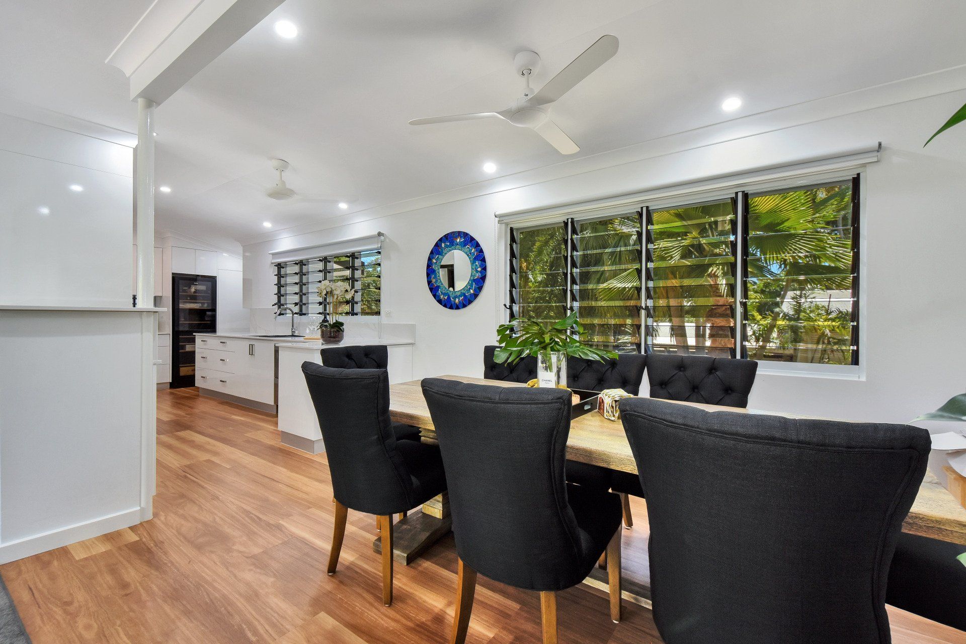 Dining room with wooden floors, dark chairs, a table, and a window with greenery visible outside.