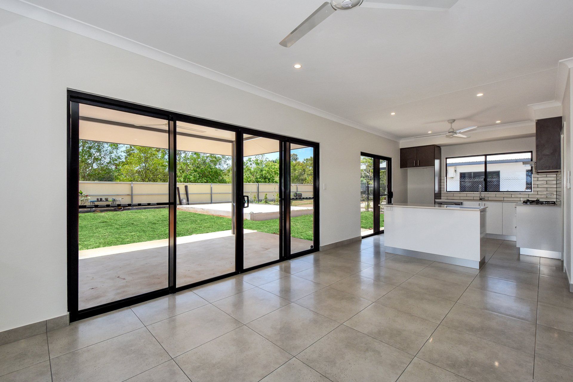 Empty, modern living room with large sliding glass doors to a backyard; kitchen with white island.