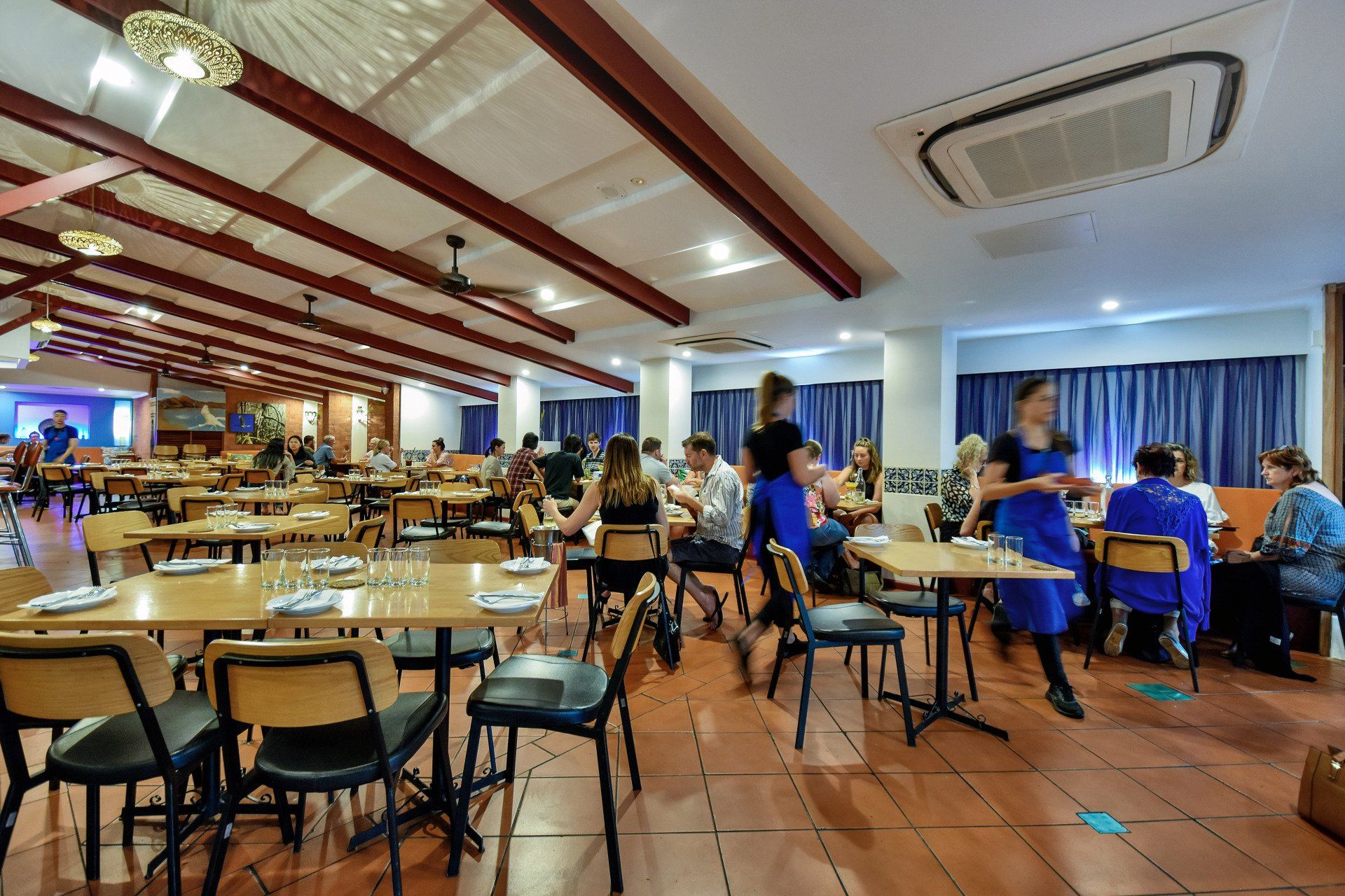 Restaurant interior with tables, chairs, and people dining. Wooden beams and tiled floors.