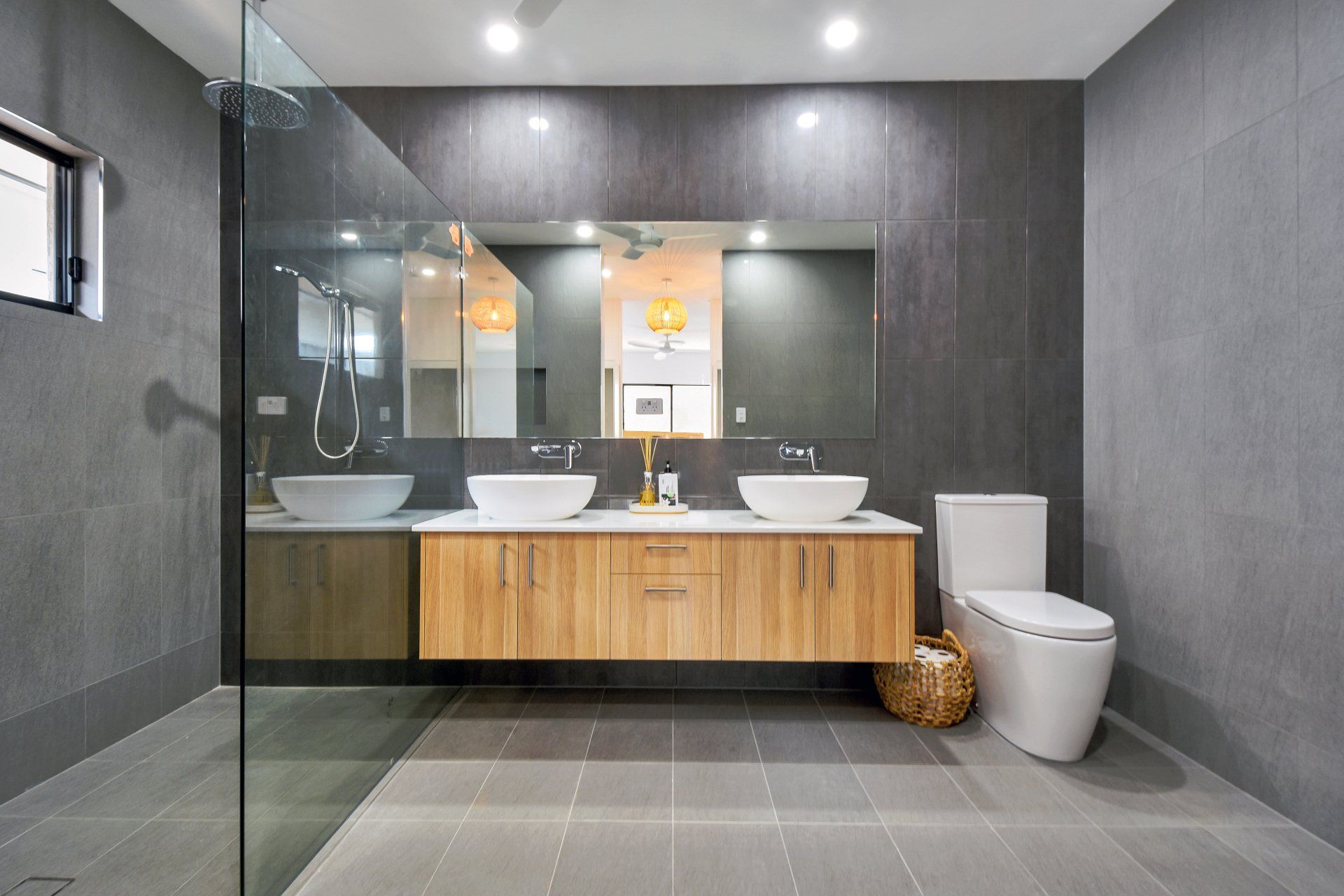 Modern gray bathroom with wood vanity, dual sinks, and toilet. Glass shower enclosure.