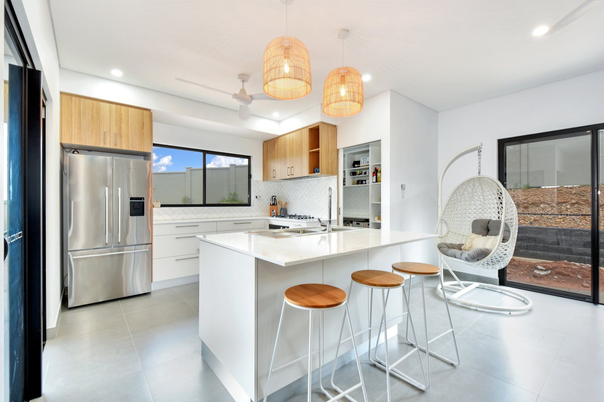 Modern kitchen with island, wooden stools, and hanging chair. Bright, white interior with wooden accents.