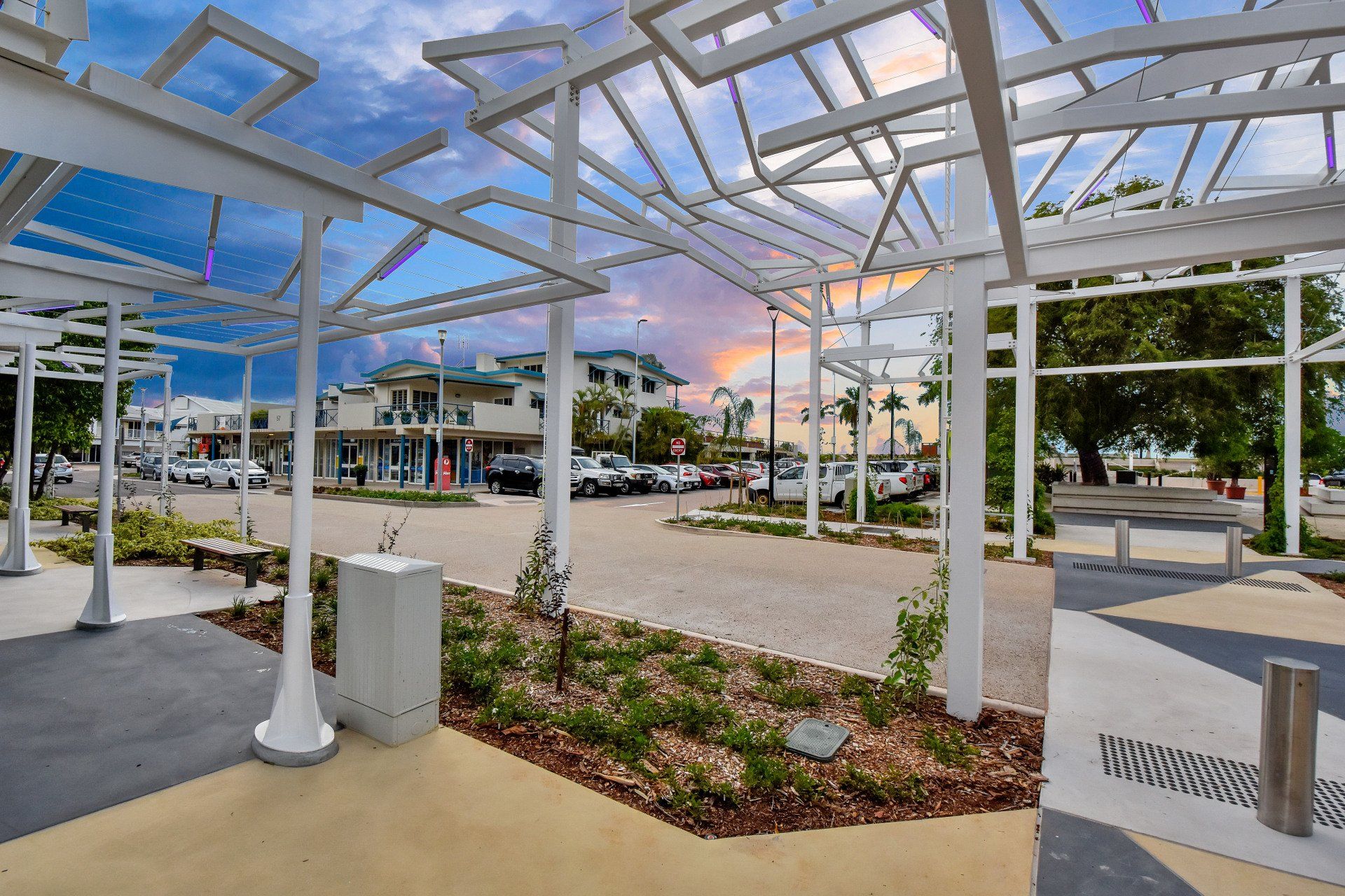 White pergola frames a street with parked cars, buildings, and a colorful sunset sky.