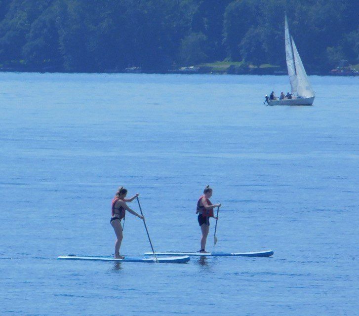 two people on paddle boards in the water with a sailboat in the background