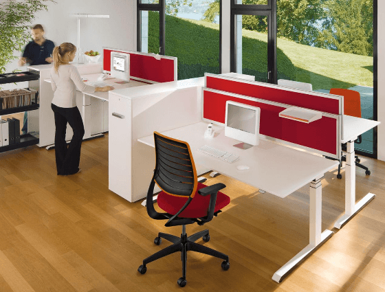 Postazione di lavoro moderna con scrivanie regolabili in altezza, schermi divisori rossi e sedie ergonomiche su un pavimento in legno.