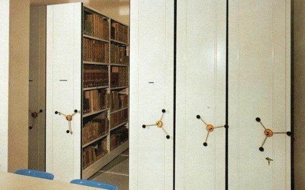 Scaffalature mobili bianche per biblioteca, con manovelle manuali, in una stanza con file di libri archiviati.