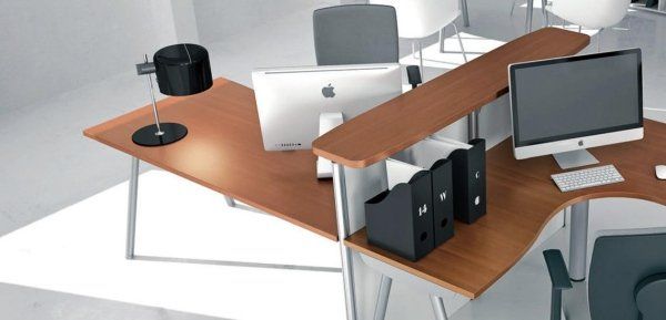 Postazione di lavoro moderna per ufficio, composta da due scrivanie in legno con computer, materiale per ufficio e sedie, in uno spazio luminoso e aperto.