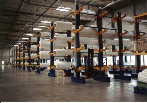 Magazzino industriale caratterizzato da alte scaffalature a sbalzo che ospitano grandi rotoli cilindrici bianchi disposti in file ordinate.