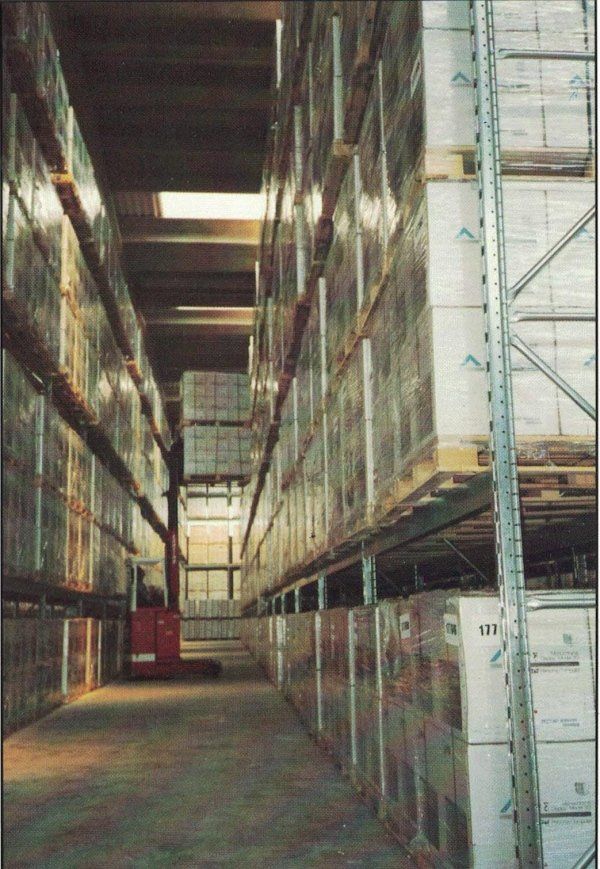 Uno stretto corridoio di un magazzino, fiancheggiato da pile di pallet di merci, con un carrello elevatore rosso in funzione in lontananza.