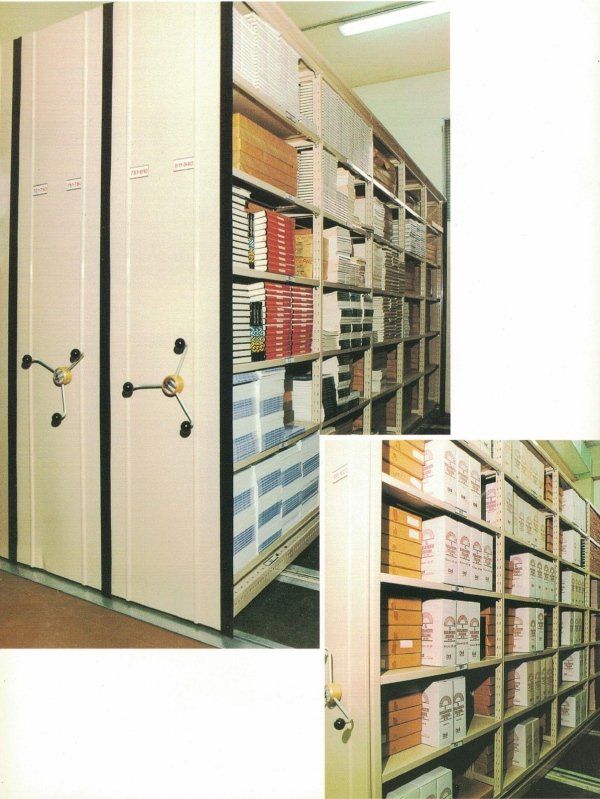 Due sistemi di archiviazione mobili ad alta densità con manovelle manuali, pieni di scatole bianche e libri su ripiani metallici.