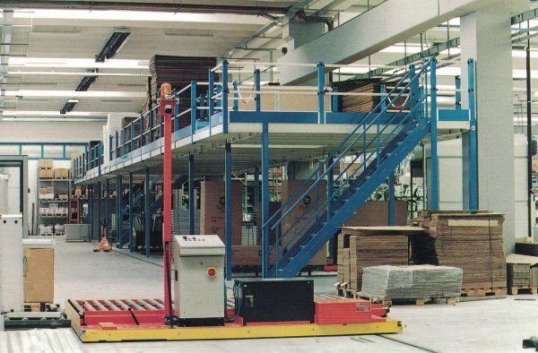 Un veicolo industriale a piattaforma di colore rosso si muove all'interno di un magazzino con una struttura a soppalco metallica blu a due piani.