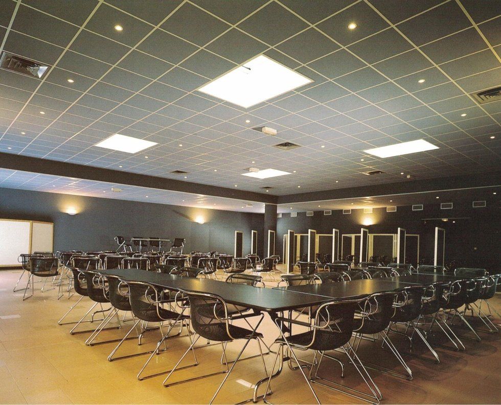 Una sala conferenze con sedie scure disposte attorno a lunghi tavoli, un soffitto a griglia con illuminazione integrata e pareti dai colori neutri.