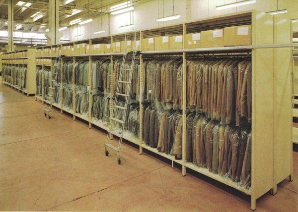 Lunghe file di abiti appesi a grucce all'interno di custodie, riposti su scaffalature industriali a più livelli in un magazzino.