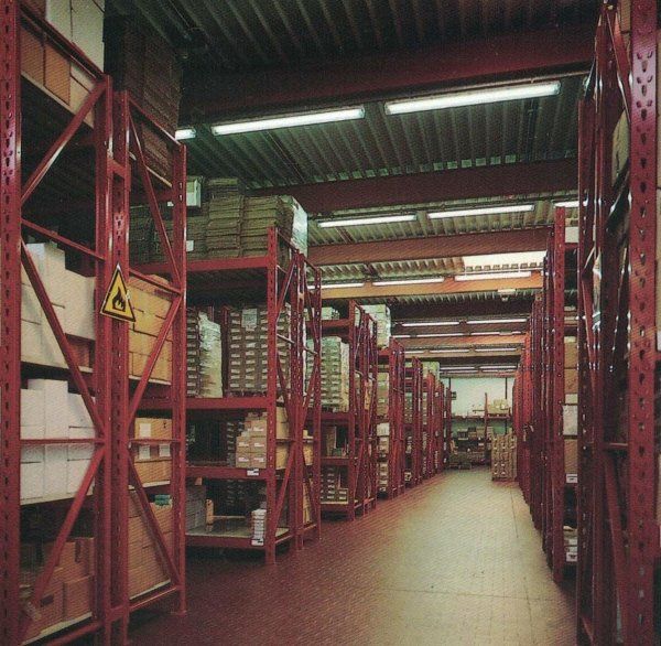 Interno di un magazzino caratterizzato da file di scaffalature industriali in metallo rosso, su cui sono appoggiate scatole di cartone su un pavimento piastrellato.