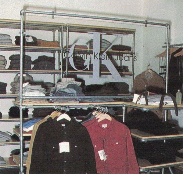 Espositore per abbigliamento con il marchio Calvin Klein Jeans, dotato di ripiani con capi piegati e camicie appese.